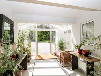 A sunlit room with glass doors, plants, a wicker sofa, tables, and a decorative screen at The River House Avon Valley Stonehenge with fishing Salisbury