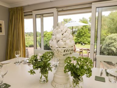 A dining table with a white ceramic fruit centerpiece and glass vases with green foliage by a sliding glass door at The River House Avon Valley Stonehenge with fishing Salisbury