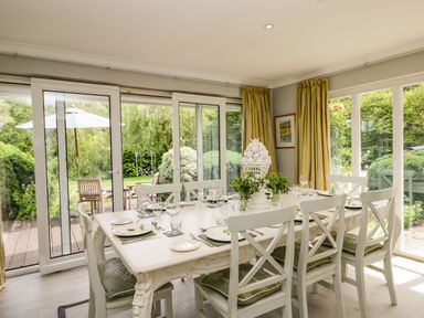 A dining room with a white table and chairs set next to large glass doors overlooking a garden at The River House Avon Valley Stonehenge with fishing Salisbury