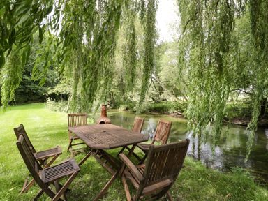 A wooden outdoor table with six wooden chairs under hanging tree branches next to a river at The River House Avon Valley Stonehenge with fishing Salisbury