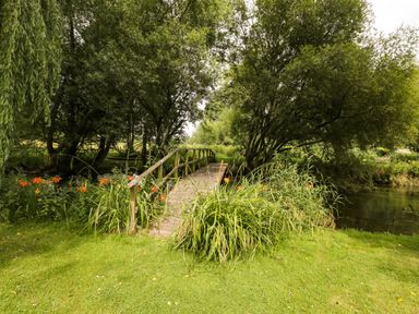 A wooden footbridge over a river surrounded by grass trees and orange flowers at The River House Avon Valley Stonehenge with fishing in Salisbury