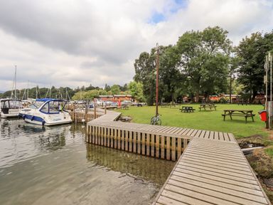 A wooden dock extending into water with boats moored beside it and picnic tables on grass near trees at Black Dog Lodge in Thirlmere 3 White Cross Bay near Windermere