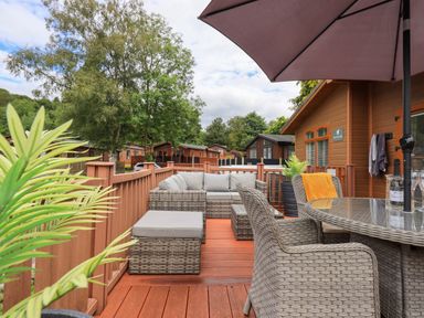 An outdoor deck with wicker furniture including chairs, a sofa, a table with an umbrella and potted plants at Black Dog Lodge in Thirlmere 3, White Cross Bay near Windermere