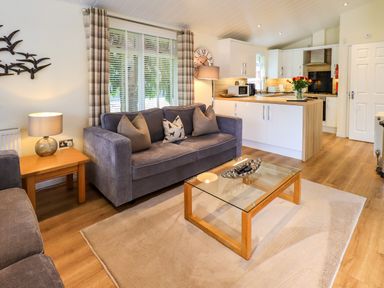 A living room with grey sofas glass coffee table wooden side table and a kitchen area at Black Dog Lodge Thirlmere 3 White Cross Bay near Windermere