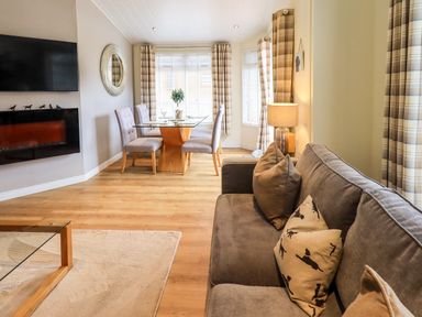 A living room with a brown sofa and cushions a glass dining table with chairs a wall-mounted TV and electric fireplace at Black Dog Lodge in Thirlmere 3 White Cross Bay near Windermere