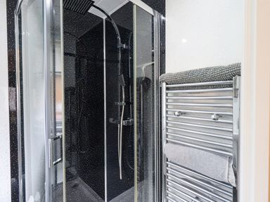 A shower with glass doors and black speckled walls next to a chrome towel radiator with folded towels at Black Dog Lodge Thirlmere 3 White Cross Bay near Windermere