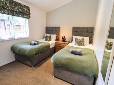A bedroom with two single beds with green throws and pillows a wooden nightstand with a lamp and a window with blinds and patterned valance at Black Dog Lodge Thirlmere 3 White Cross Bay near Windermere
