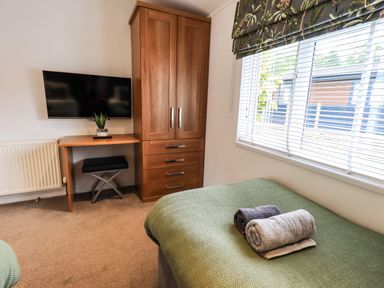 A bedroom with a green bedspread two rolled towels a wooden wardrobe a desk a stool a wall-mounted television and a window with blinds and patterned valance at Black Dog Lodge Thirlmere 3 White Cross Bay near Windermere