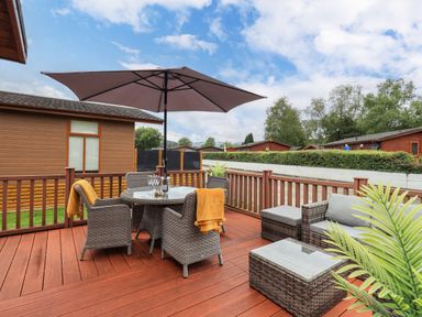 An outdoor patio with wicker furniture a round dining table with an umbrella and nearby houses and trees at Black Dog Lodge Thirlmere 3 White Cross Bay near Windermere