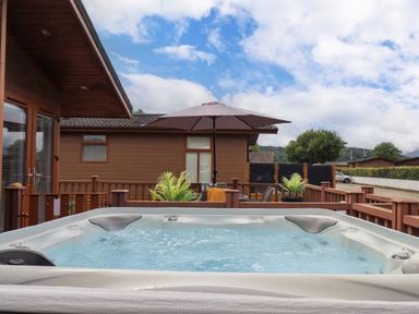 An outdoor hot tub on a wooden deck with a patio umbrella and lounge chairs at Black Dog Lodge Thirlmere 3 White Cross Bay near Windermere