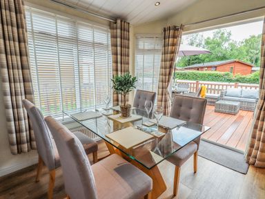 A dining area with a glass table and four chairs next to large windows and an open door leading to a patio with outdoor furniture at Black Dog Lodge Thirlmere 3 White Cross Bay near Windermere