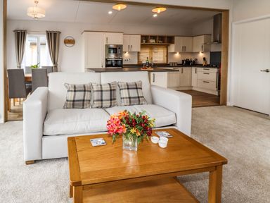 A living area with a white sofa and plaid cushions and a wooden coffee table with flowers with an open kitchen in the background at Bramblewick in Runswick Bay near Staithes