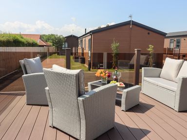 An outdoor seating area with wicker chairs a loveseat a glass table with flowers and drinks on a wooden deck at Bramblewick in Runswick Bay near Staithes