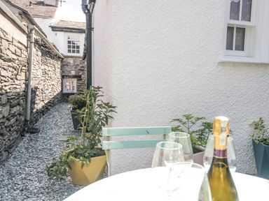 A narrow outdoor space with stone walls and potted plants alongside a white wall with a window and a table with glasses and a bottle at Old Toffee Shop Ambleside