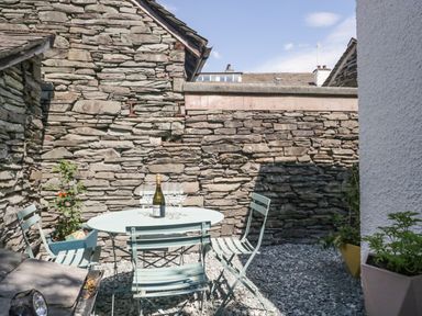 A small outdoor seating area with a round table four chairs a bottle and glasses on the table stone walls and plants in pots at Old Toffee Shop in Ambleside
