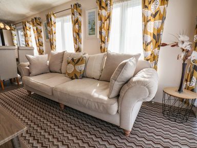 A beige sofa with cushions next to windows with patterned curtains and a small round side table with flowers at The Rodley Pines in Cayton