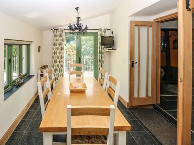 A dining room with a wooden table and six chairs near green windows and a glass door with curtains at Trewrach Cottage in Dinas Cross