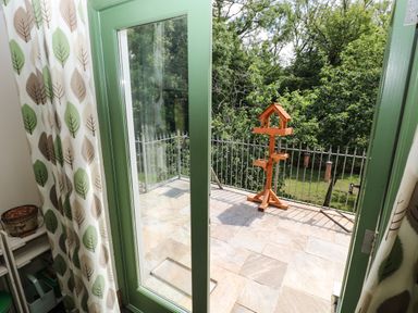 A view through green framed glass doors onto a stone tiled balcony with a wooden bird feeder and metal railing surrounded by trees at Trewrach Cottage in Dinas Cross