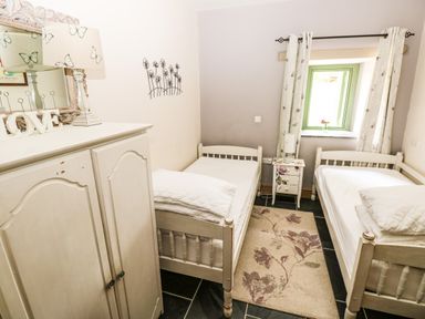 A bedroom with two single beds a small nightstand between them a cabinet with a lamp and decorative mirror and a window with curtains at Trewrach Cottage in Dinas Cross