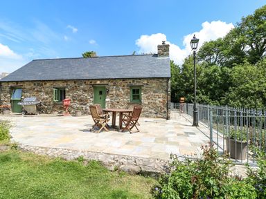 A stone cottage with slate roof and green doors and windows a patio with wooden table and chairs barbecue and railing with lamp post outside at Trewrach Cottage in Dinas Cross