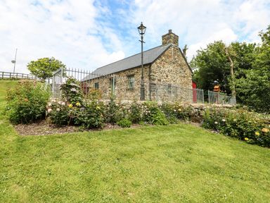 A stone cottage with a slate roof behind a metal fence surrounded by flower beds and a grassy lawn at Trewrach Cottage in Dinas Cross