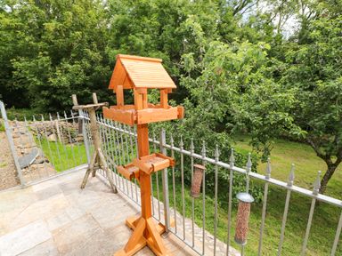 A patio with a wooden bird feeder and metal railings overlooking a green garden with trees at Trewrach Cottage in Dinas Cross