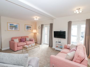 A living room with two pink sofas a grey armchair a glass coffee table a wall-mounted TV and two sets of French doors with curtains at Weydale Avenue in Scarborough