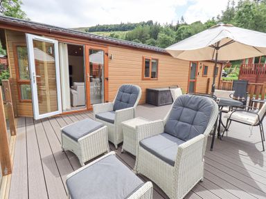An outdoor deck with two armchairs and footrests a dining table with chairs and a large umbrella at Esthwaite Lodge Beckside 43 Limefitt Park near Windermere