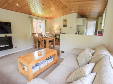 A living room with a beige sofa wooden coffee table dining table with chairs and a television on the wall at Esthwaite Lodge Beckside 43 Limefitt Park near Windermere