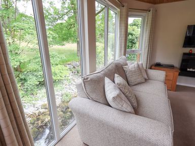 A living room with a beige sofa and cushions next to large windows showing greenery at Esthwaite Lodge in Beckside 43 Limefitt Park near Windermere