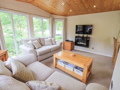 A living room with two sofas a wooden coffee table a wall-mounted TV and large windows at Esthwaite Lodge Beckside 43 Limefitt Park near Windermere