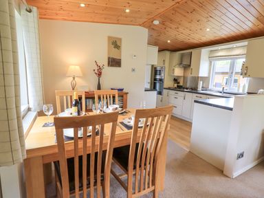 A dining area with a wooden table set for four and a kitchen with white cabinets at Esthwaite Lodge Beckside 43 Limefitt Park near Windermere