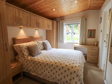 A bedroom with a bed under wooden cabinets a window with curtains and a wooden desk with drawers at Esthwaite Lodge Beckside 43 Limefitt Park near Windermere