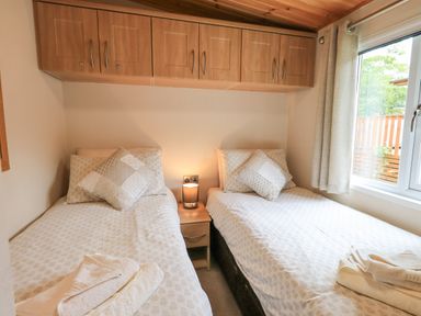 A bedroom with two single beds a nightstand with a lamp and overhead cabinets at Esthwaite Lodge Beckside 43 Limefitt Park near Windermere