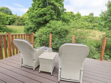 A balcony with two wicker chairs and a wicker table overlooking green trees and grass at Esthwaite Lodge in Beckside 43 Limefitt Park near Windermere