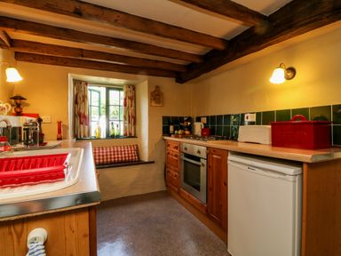 A kitchen with wooden cabinets a white fridge an oven a window with a seat below and red kitchen accessories at Bell Buoy Cottage in Morwenstow