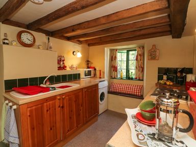 A kitchen with wooden cabinets a sink a microwave a clock and a window seat at Bell Buoy Cottage in Morwenstow