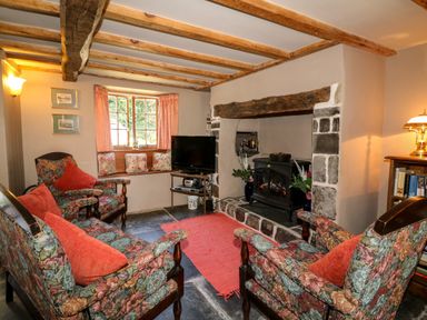 A sitting room with floral armchairs a wood stove on a stone hearth a television and bookshelf at Bell Buoy Cottage Morwenstow
