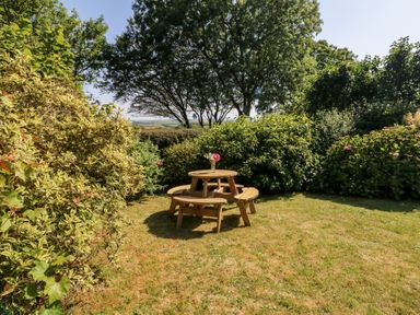 A garden with a round wooden picnic table and benches surrounded by bushes and trees at Bell Buoy Cottage in Morwenstow