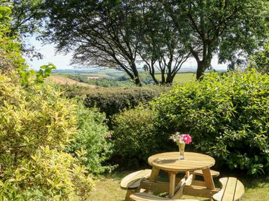 A wooden picnic table with a flower vase surrounded by bushes and trees in a garden with a countryside view at Bell Buoy Cottage in Morwenstow