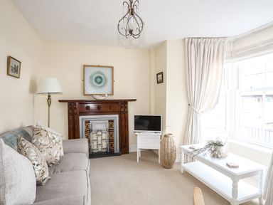 A living room with a sofa, wooden fireplace, television, floor lamp, white coffee table, and large window with curtains at Hyfrydle in Borth-Y-Gest