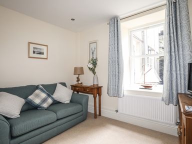 A living room with a blue sofa and cushions a wooden side table with a lamp and vase a window with blue curtains and a model sailboat on the sill at Hyfrydle in Borth-Y-Gest