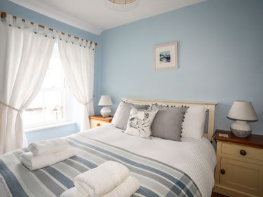 A bedroom with a bed with pillows and folded towels on a striped blanket beside two bedside tables with lamps and a window with curtains at Hyfrydle in Borth-Y-Gest