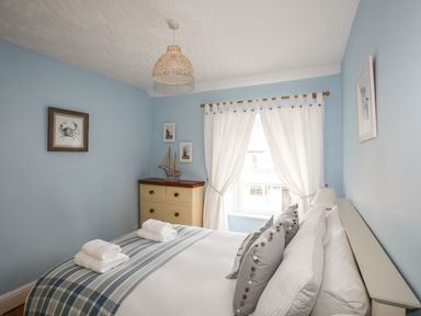A bedroom with blue walls a bed with pillows and folded towels a chest of drawers and a window with white curtains at Hyfrydle in Borth-Y-Gest