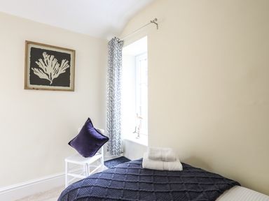 A bedroom with a bed covered by a dark blanket and folded towels on it a white chair with a purple cushion a window with patterned curtains and artwork on the wall at Hyfrydle in Borth-Y-Gest