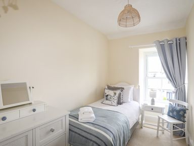 A bedroom with a single bed next to a window with blue curtains a bedside table a chair with a pillow and a white dresser with a mirror at Hyfrydle in Borth-Y-Gest