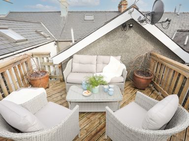 An outdoor wooden deck with wicker sofa and chairs around a glass coffee table with plants and cups at Hyfrydle in Borth-Y-Gest
