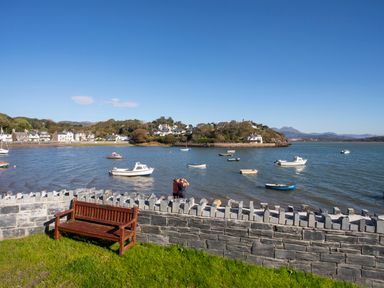 A wooden bench on grass with a stone wall overlooking boats on water and houses in the background at Hyfrydle in Borth-Y-Gest