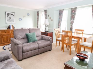 A living room with a grey sofa a wooden dining table with chairs and floral curtains at Ellan Vannin in Sheringham