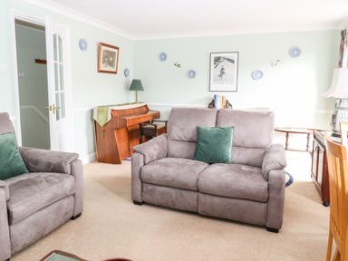 A living room with gray sofas a wooden piano a table lamp and framed pictures on the wall at Ellan Vannin in Sheringham